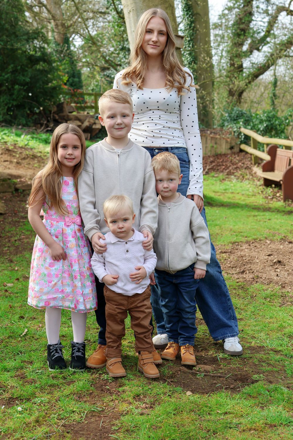 A young woman poses outdoors with four children of varying ages in a lush green park setting.