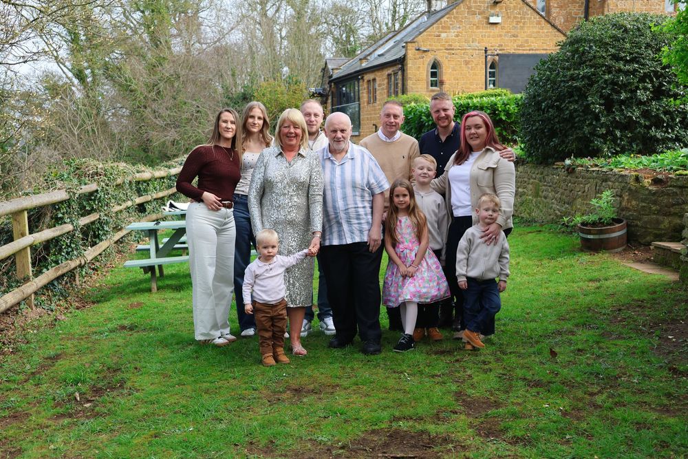 Large multigenerational family group smiling together in a lush green garden beside a wooden fence.
