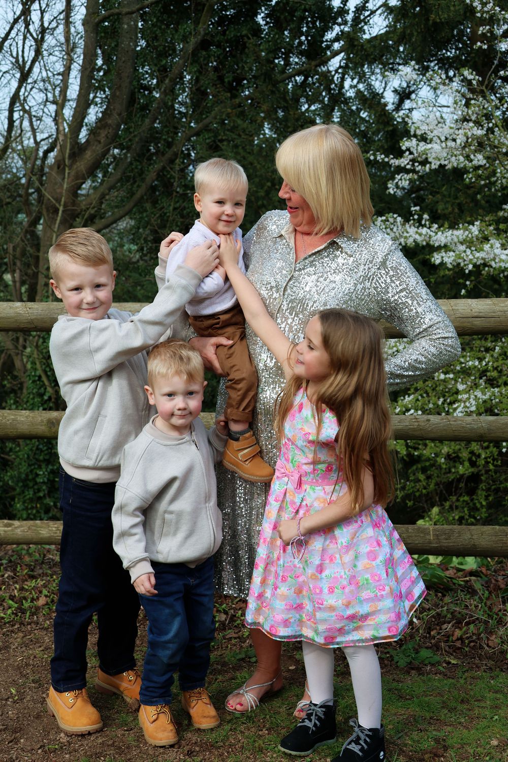 Grandmother in silver sequin dress lovingly holding four young grandchildren by a wooden fence outdoors.