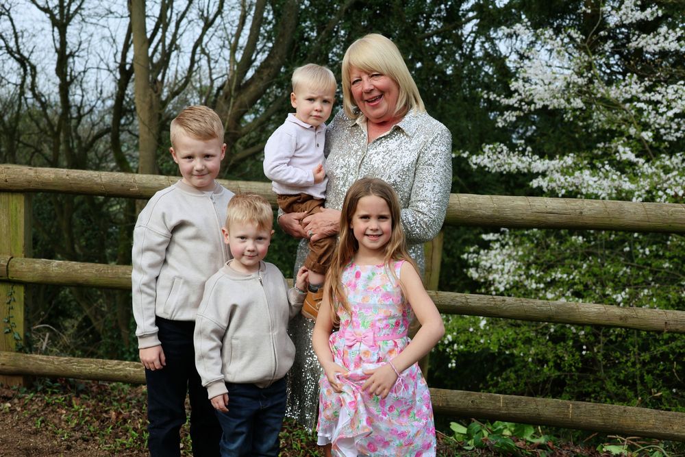 Grandmother smiling outdoors with four young children near a wooden fence, surrounded by spring trees in bloom.