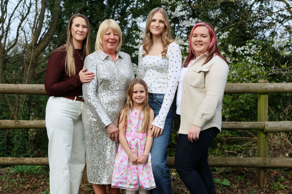 Five women and girls of different ages pose together outdoors near a wooden fence surrounded by greenery.