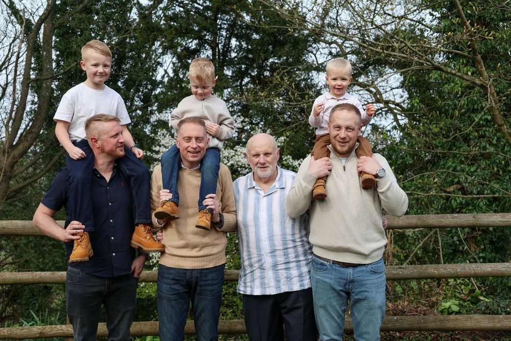 Three generations of men smile outdoors, with young children sitting on shoulders near a wooden fence surrounded by trees.