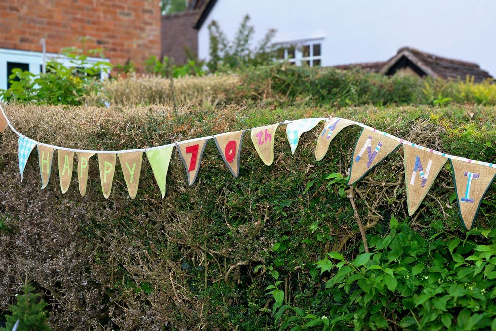 Colorful fabric bunting flags strung along a green hedge in an English garden on an overcast day.
