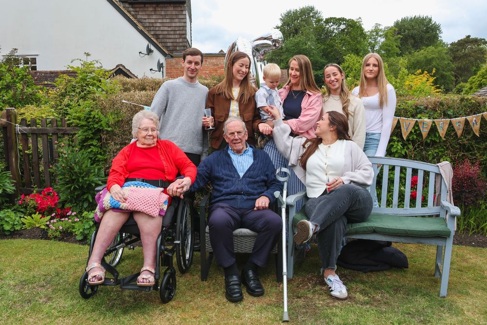 Multi-generational family gathering outdoors in a garden, including elderly members in wheelchairs and young adults smiling together.