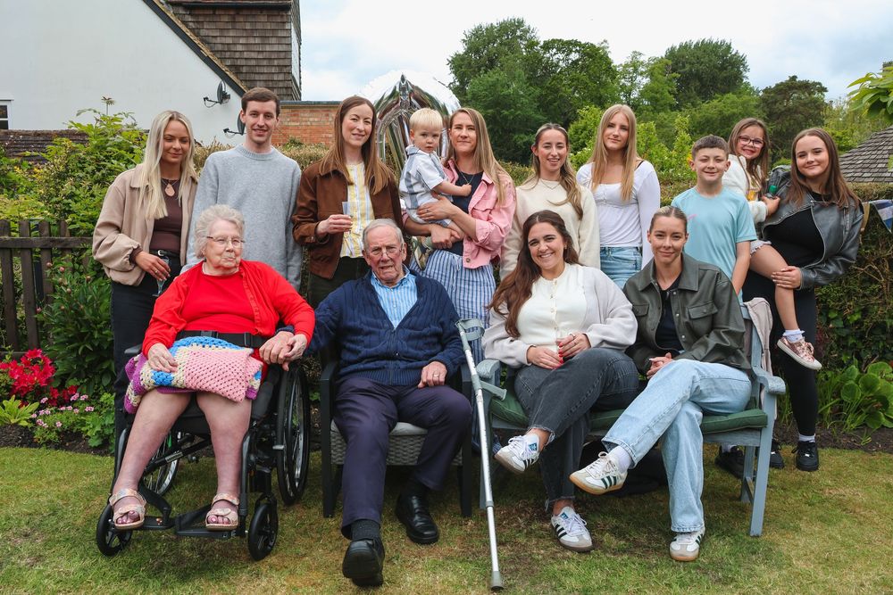 Multi-generational family group portrait outdoors in garden, ranging from elderly couple in wheelchairs to young adults and teenagers.