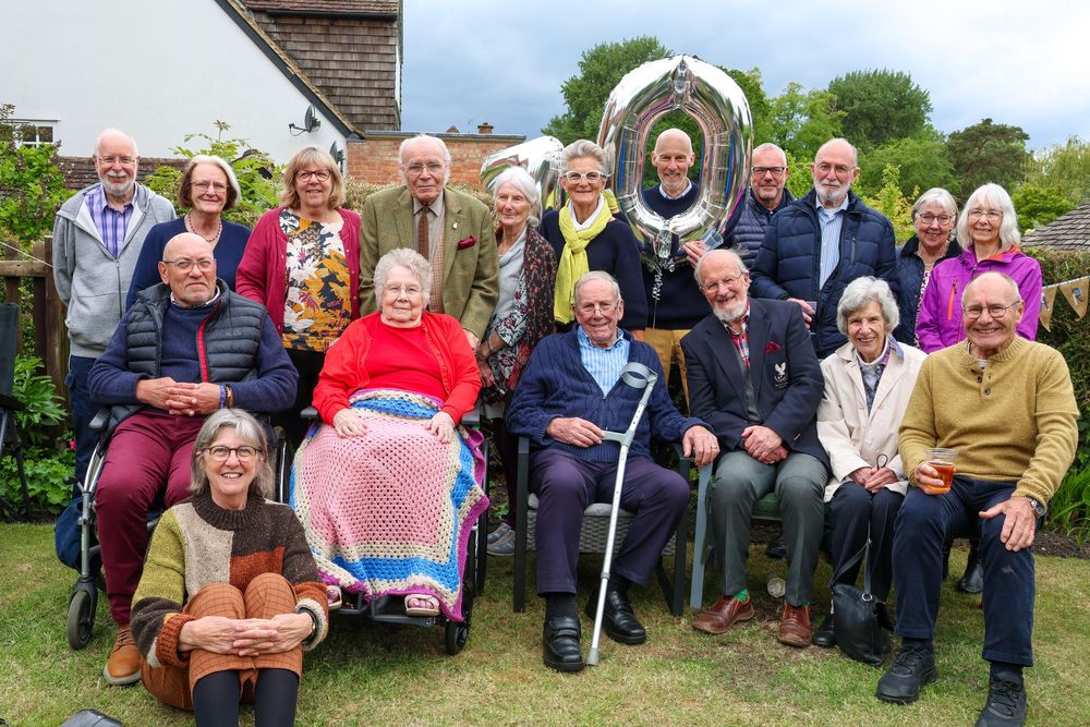 Large group of elderly people posing together outdoors in a garden, seated and standing, smiling for a group photo.