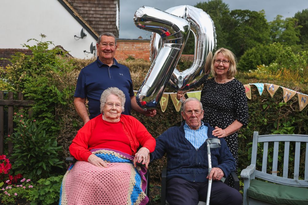 Elderly couple seated outdoors celebrating 75th anniversary with family holding silver balloon numbers in a garden.