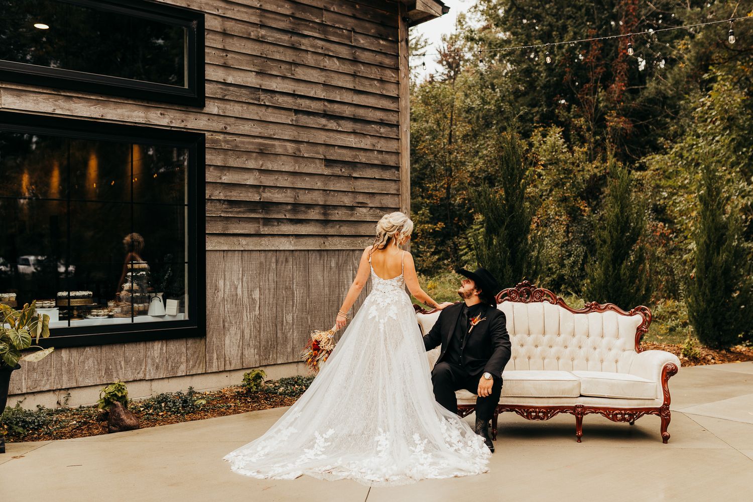 A wedding couple sits together on an elegant white sofa outdoors against a rustic wooden building backdrop.