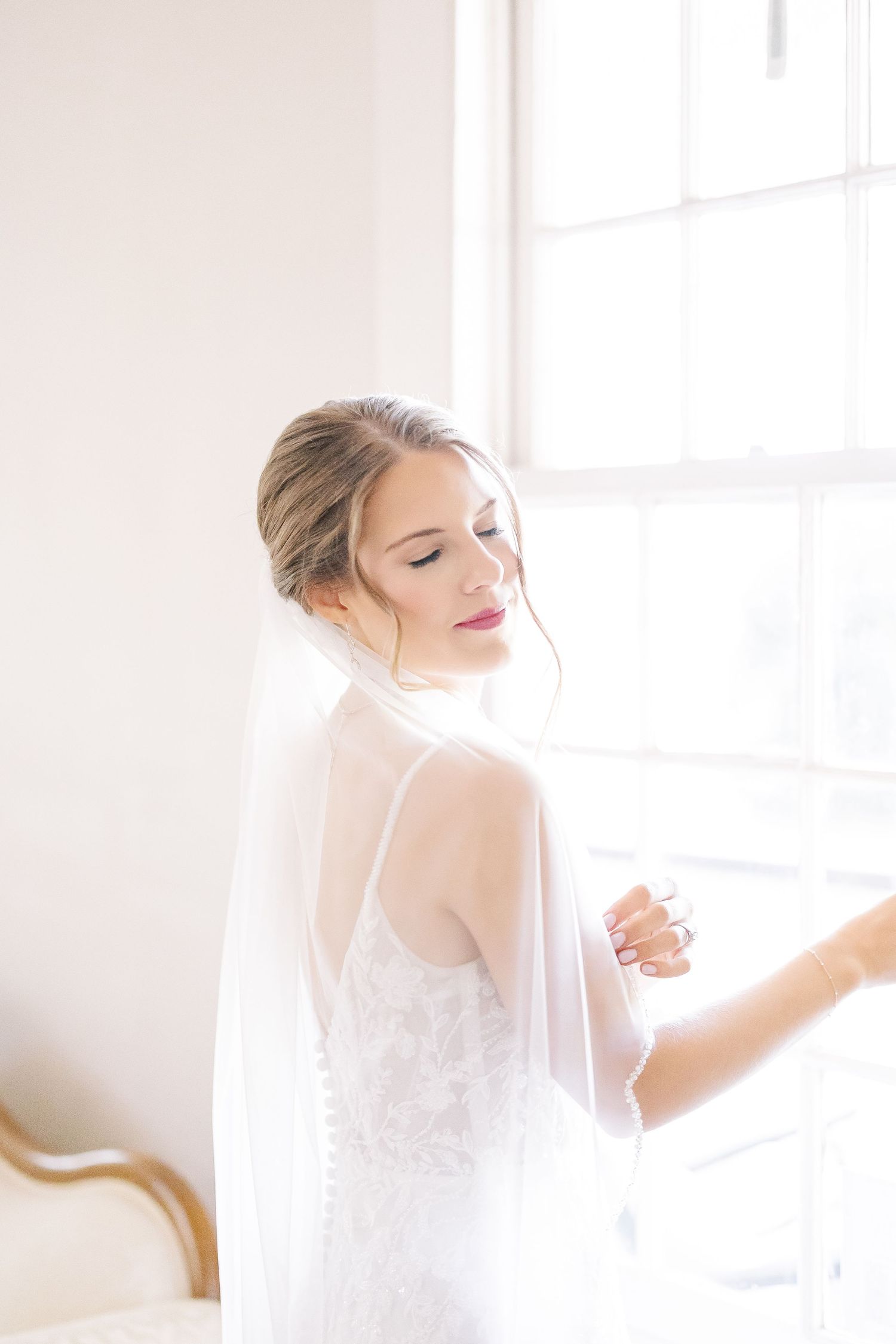 A bride in a delicate white lace wedding dress and veil is captured in a bright, sunlit room.