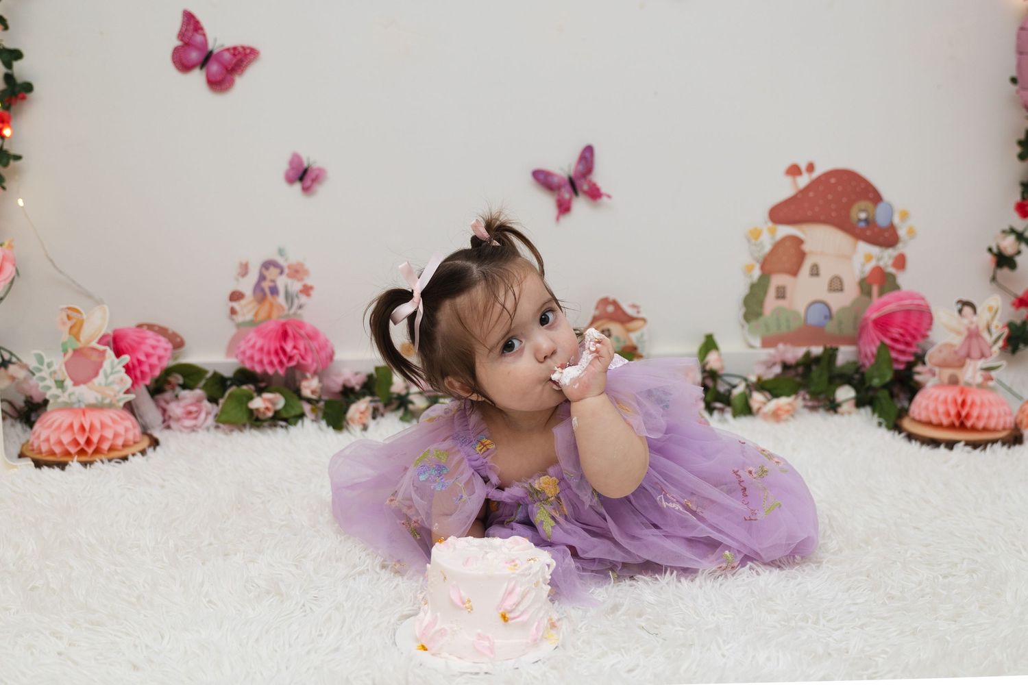 A baby in a purple tutu dress poses on a white backdrop with pink butterflies and floral decorations for a photoshoot.