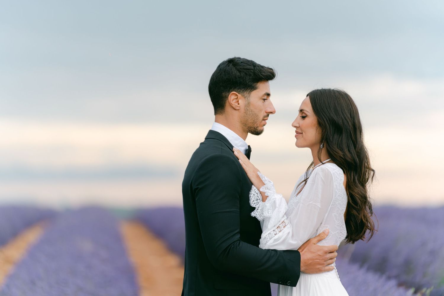 A bride in a flowing white lace wedding dress walks gracefully along a pristine beach at sunset.
