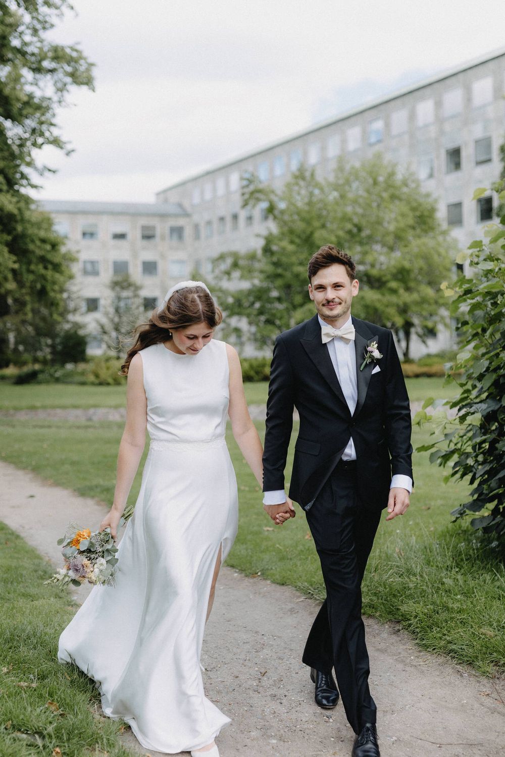 A couple in wedding attire walks along a path surrounded by greenery and buildings on a cloudy day.