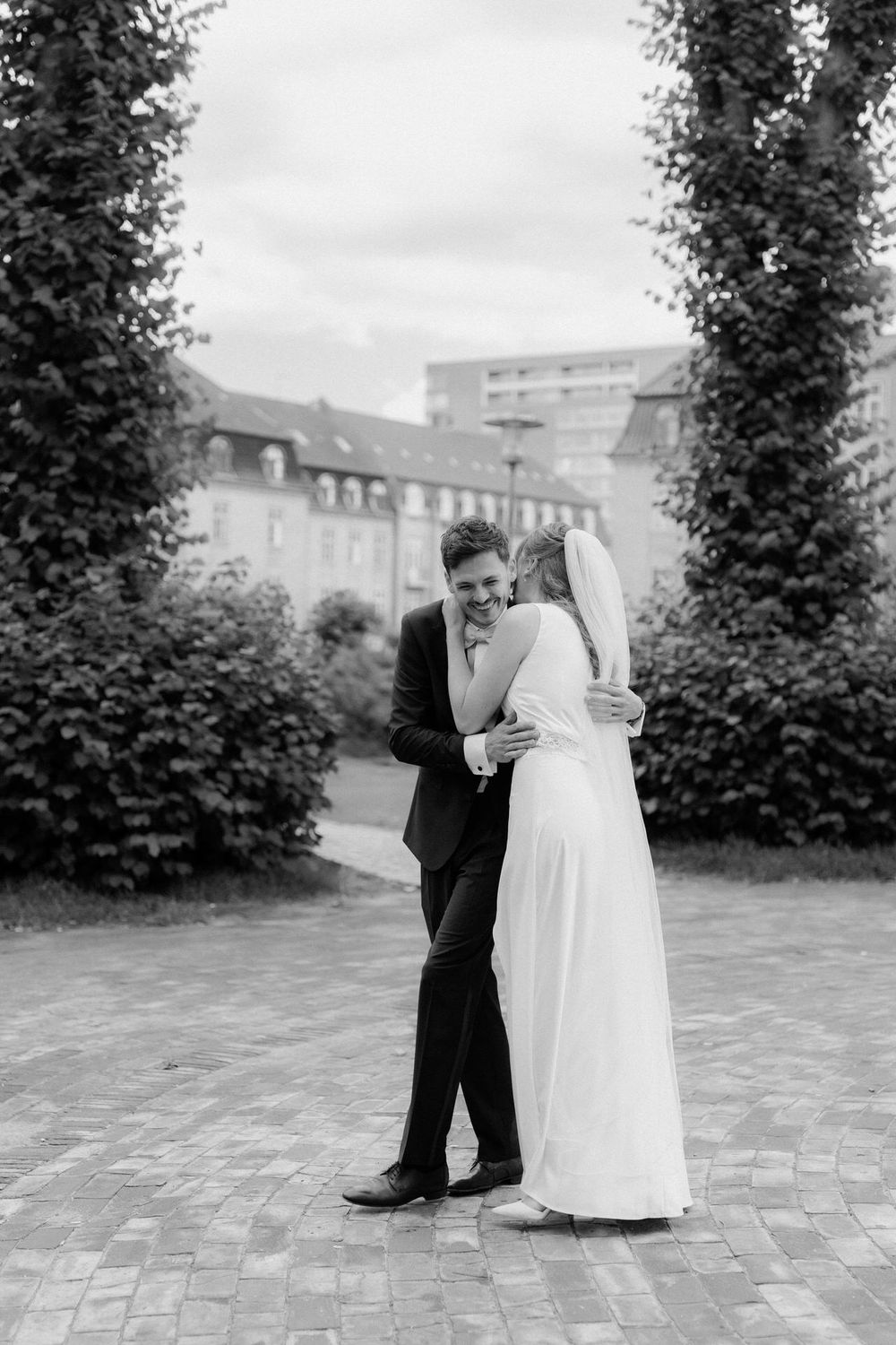A wedding couple shares intimate moments on a brick paved courtyard surrounded by tall greenery.