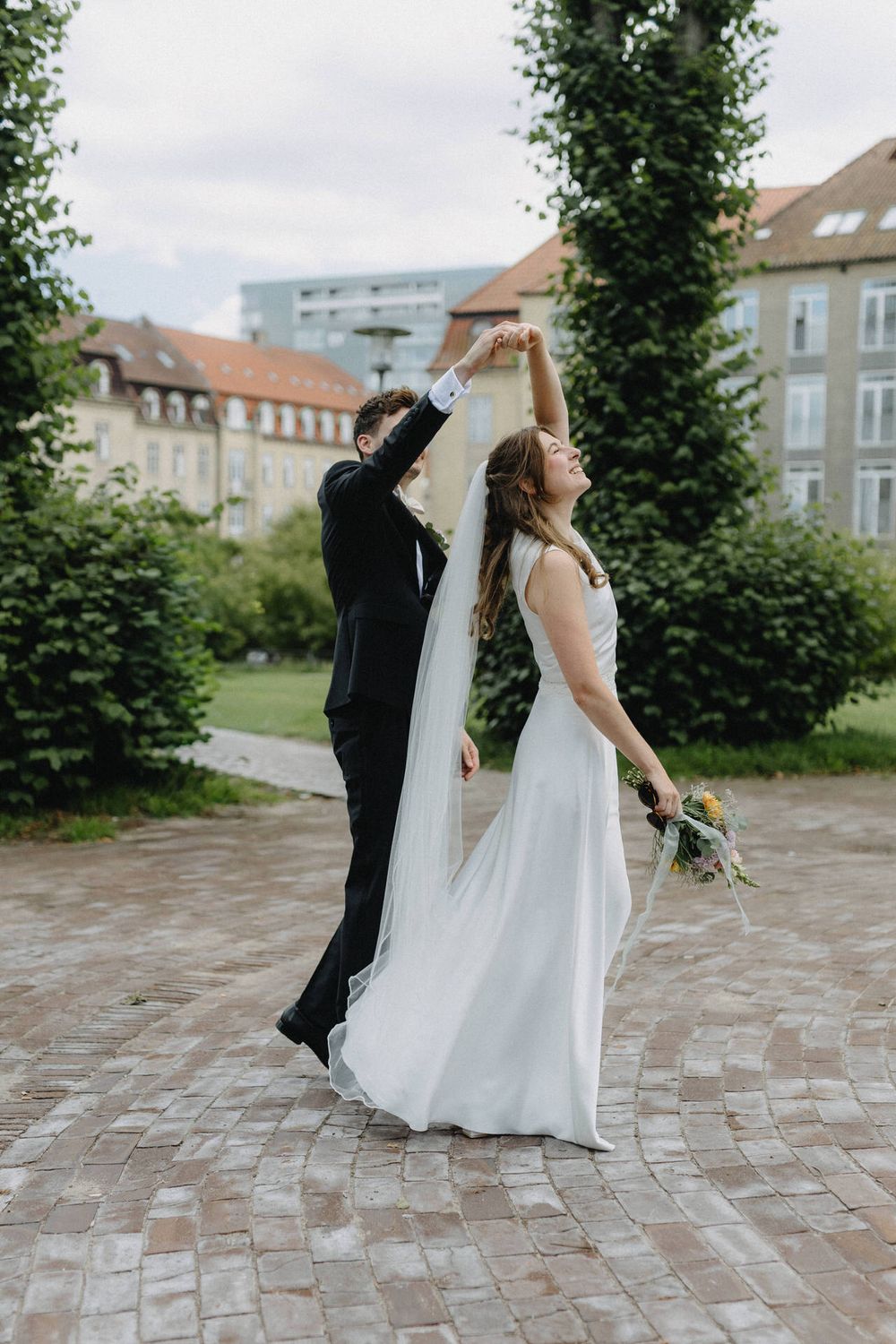 A playful wedding couple dances and twirls together on a brick patio with evergreen trees in the background.