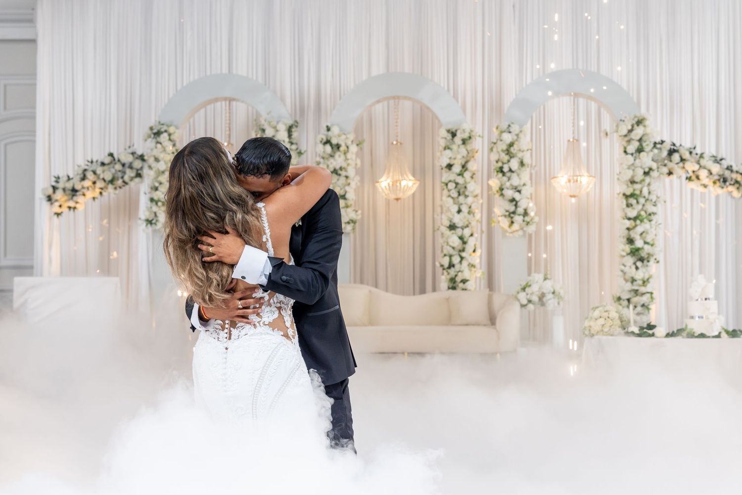 A romantic wedding couple embraces during their first dance in an elegant white and gold decorated ballroom.