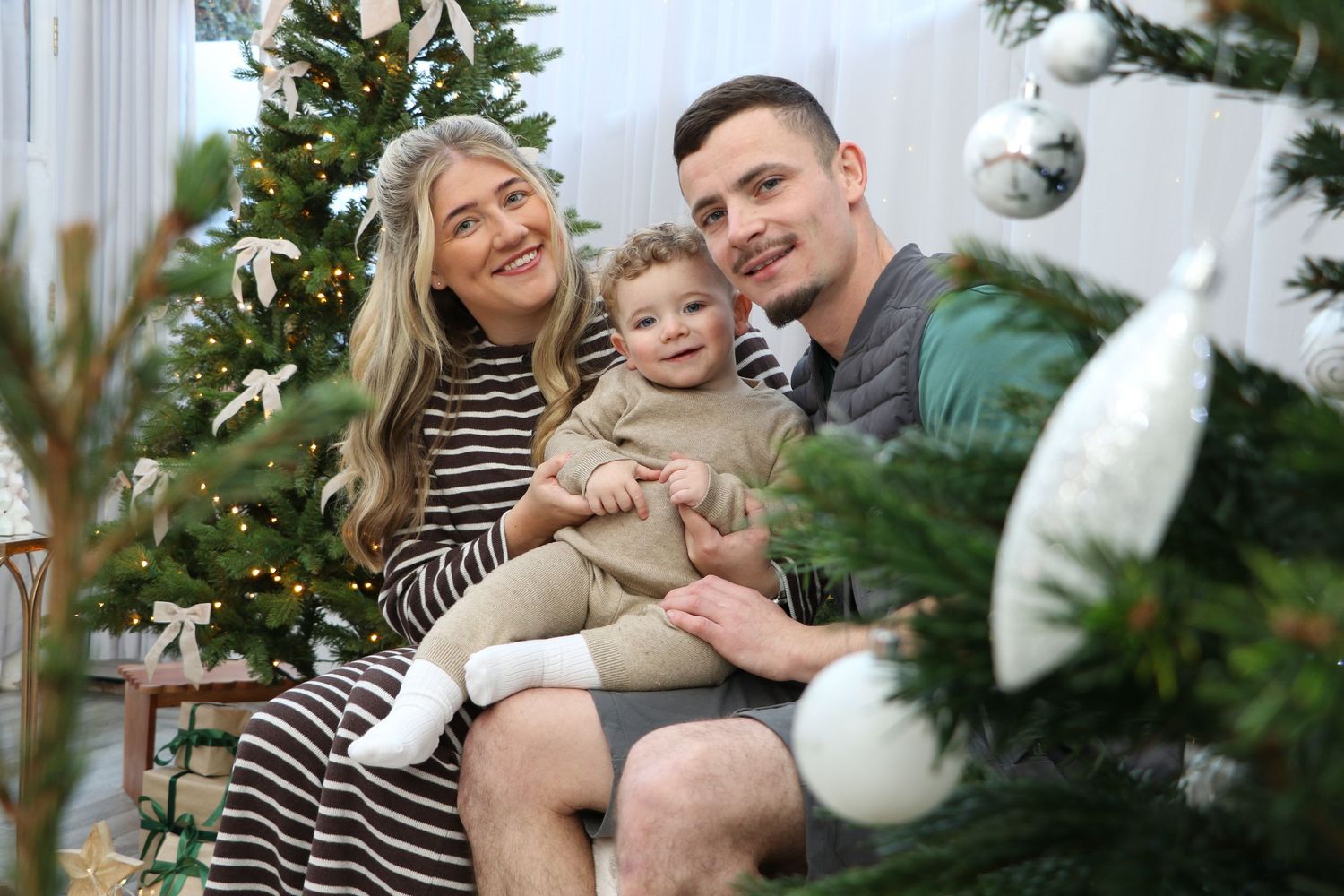 Family laughs and embraces while sitting together in front of a decorated Christmas tree during holiday photoshoot.