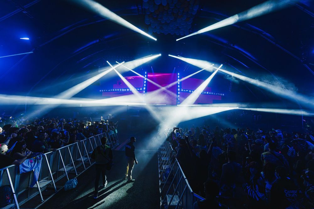Dramatic white spotlights and laser beams illuminate a dark concert venue filled with spectators behind crowd barriers.