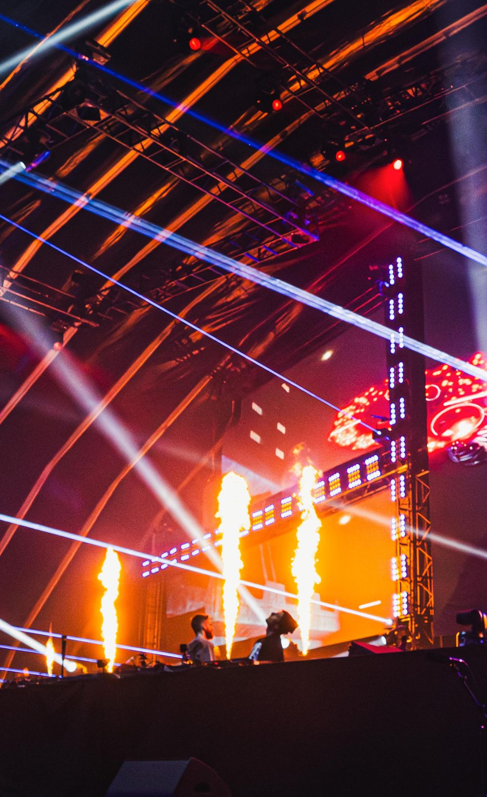 Dramatic concert stage with vibrant orange flames and blue laser lights creating an immersive atmosphere during performance.