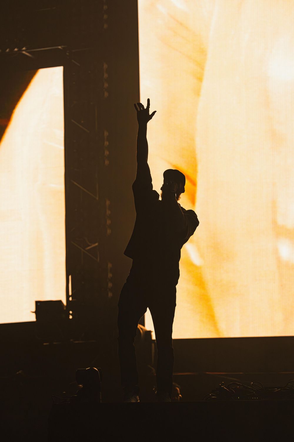 Silhouetted figures on stage perform against a bright orange and yellow backlit backdrop during a concert performance.