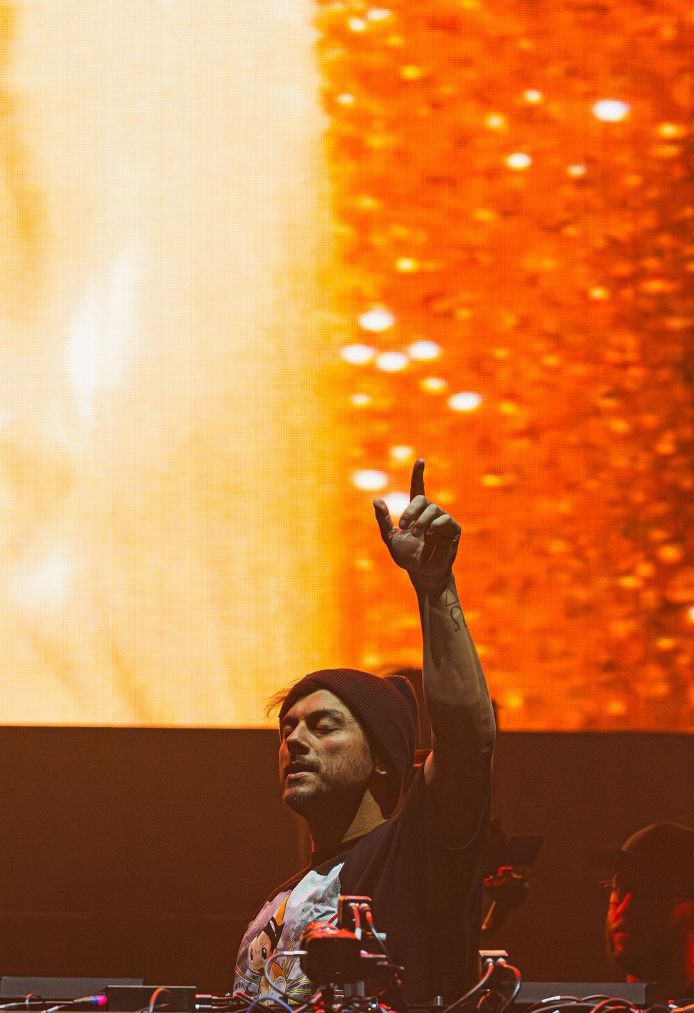DJ performs at a concert with raised hand against vibrant orange lighting and backdrop.