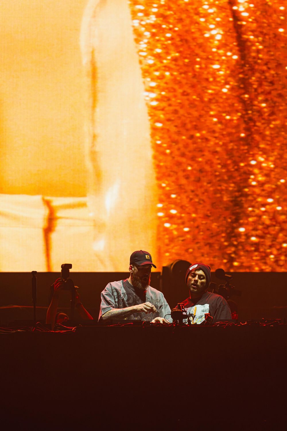 DJs perform on stage with vibrant orange lighting and glittering effects creating an energetic concert atmosphere.