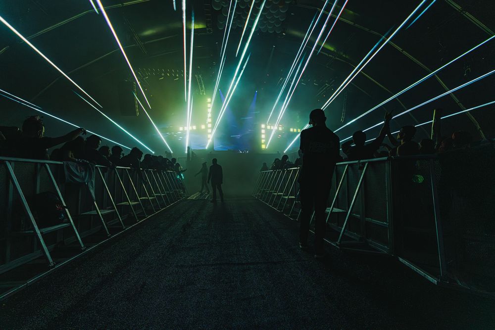Silhouettes of people walking through a futuristic tunnel with green laser lights and fog effects.