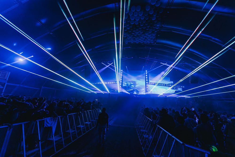 Blue laser beams illuminate a dark concert venue with silhouetted crowd and empty chairs in the foreground.