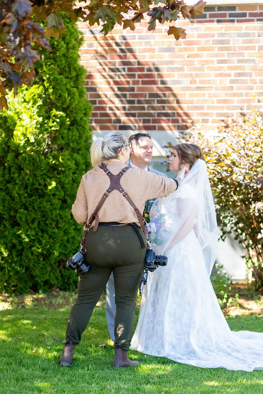 A photographer in suspenders captures a bride in a flowing white gown on a sunny day against a brick wall.