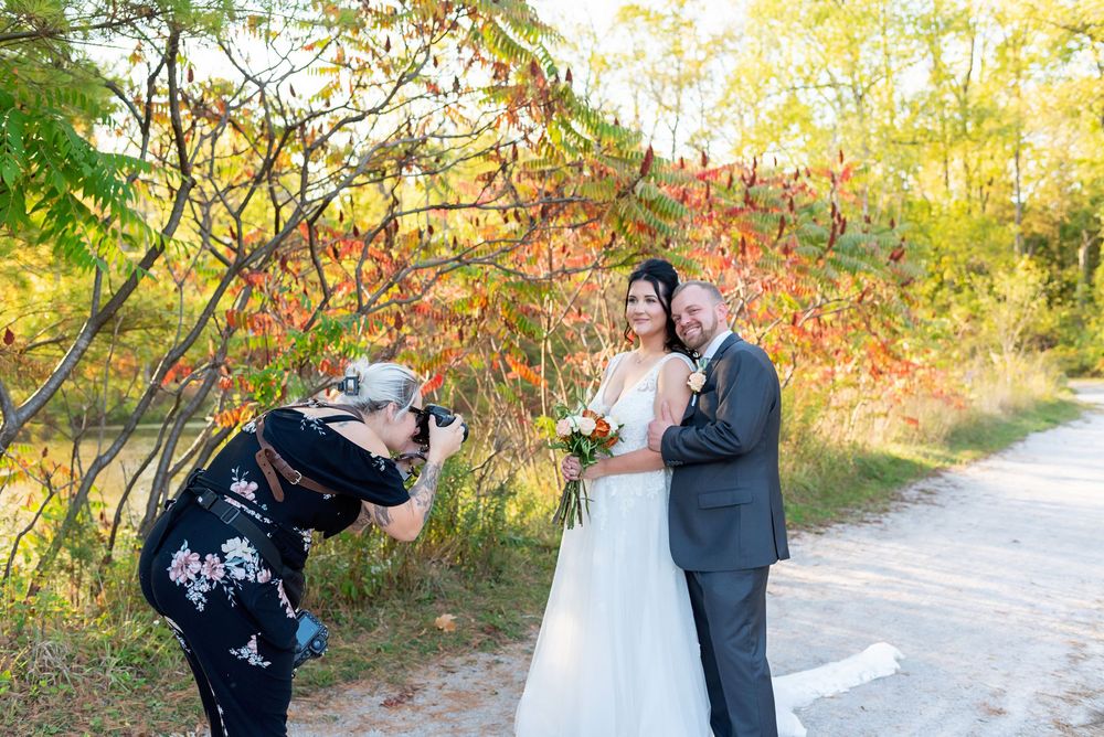 A couple in wedding attire poses together in a scenic autumn setting with golden foliage in the background.