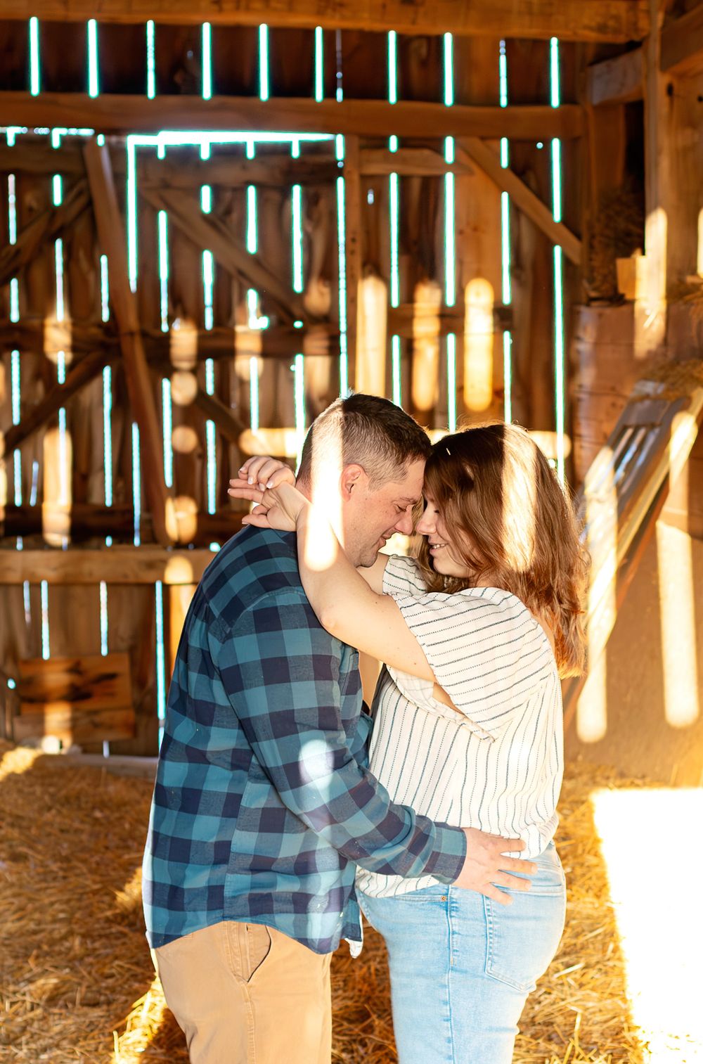 Romantic couple embraces in rustic barn with sunlight streaming through wooden slats creating magical atmosphere.