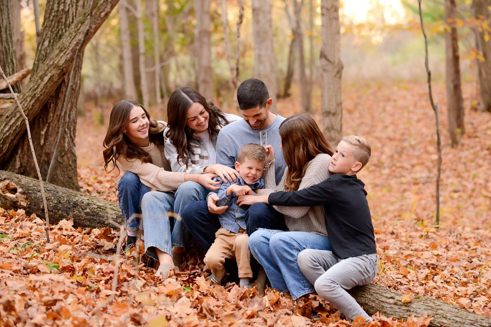 Family wearing blue and gray sits together on autumn leaves during outdoor photo session in wooded area  AT KOMOKA PROVINCIAL PARK.