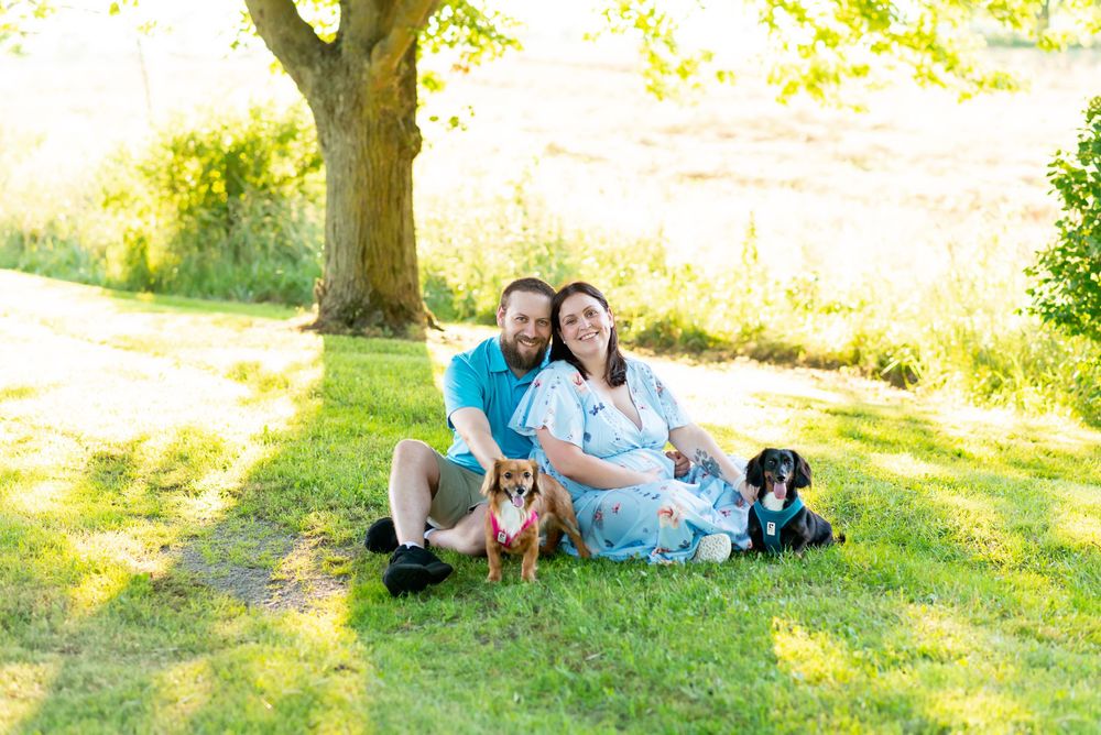 A black dog sits with people on a grassy lawn during a sunny outdoor photo session in Strathroy ontario.