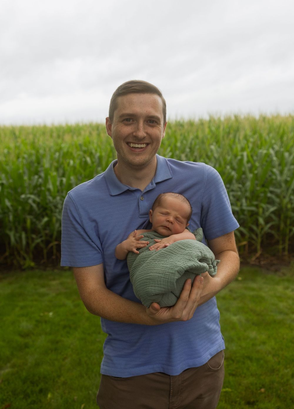 Parent in blue shirt holding newborn baby outdoors with cornfield in background.