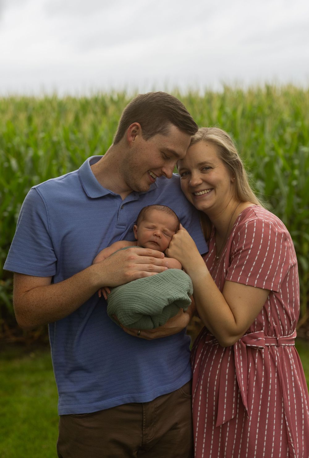 Family portrait outdoors with cornfield backdrop showing tender moment with newborn.