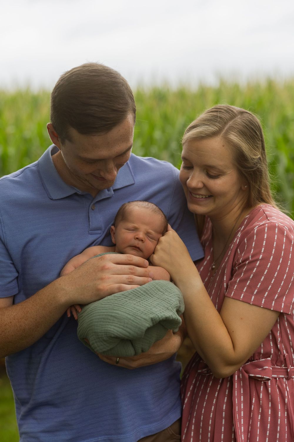 Parents sharing intimate moment with newborn baby in front of cornfield.