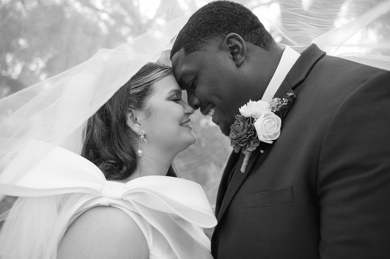 Black and white wedding photo showing tender moment between newlyweds in traditional attire with veil and boutonniere.