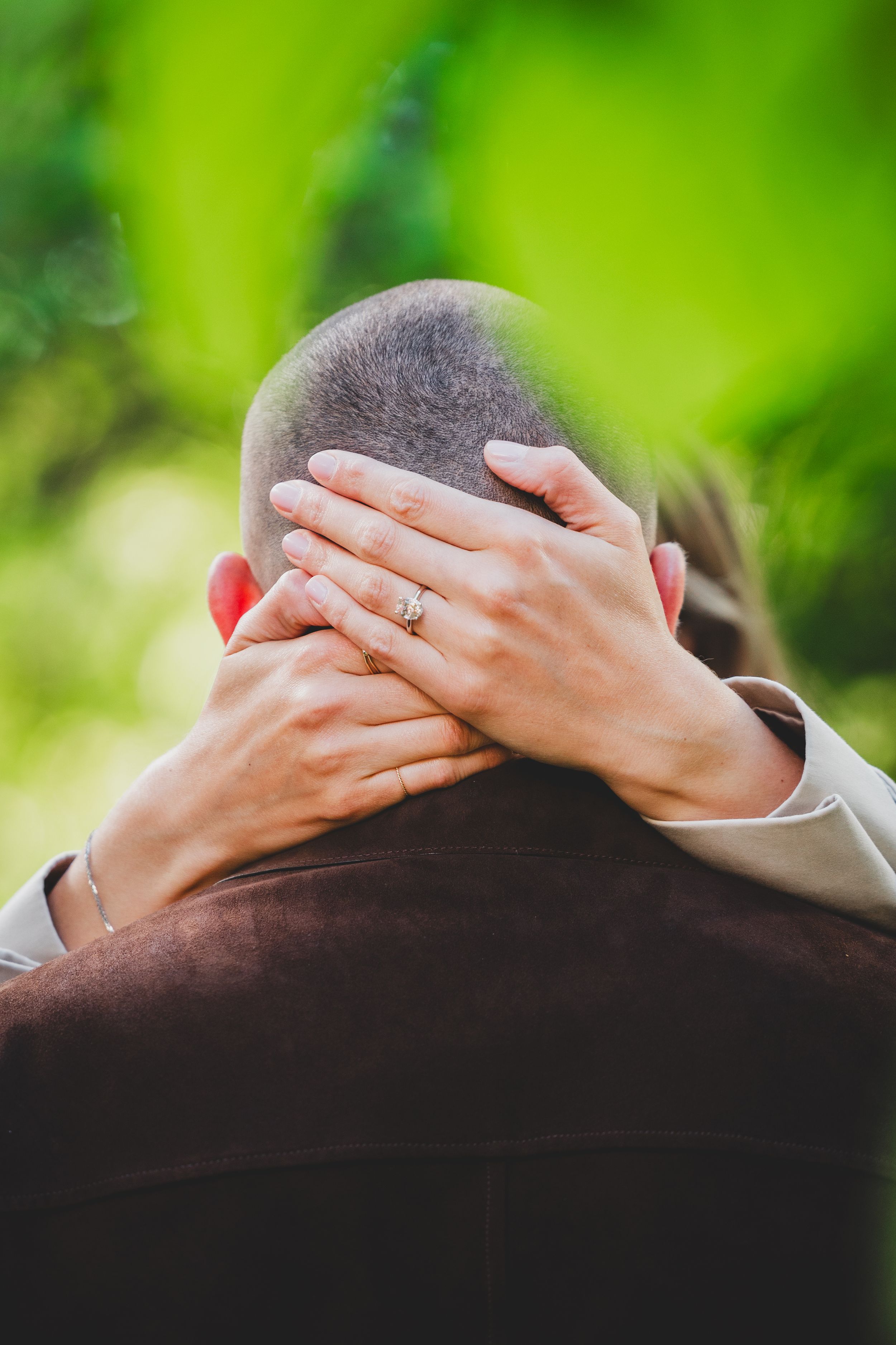 A close-up view of hands with an engagement ring against a blurred green natural background.