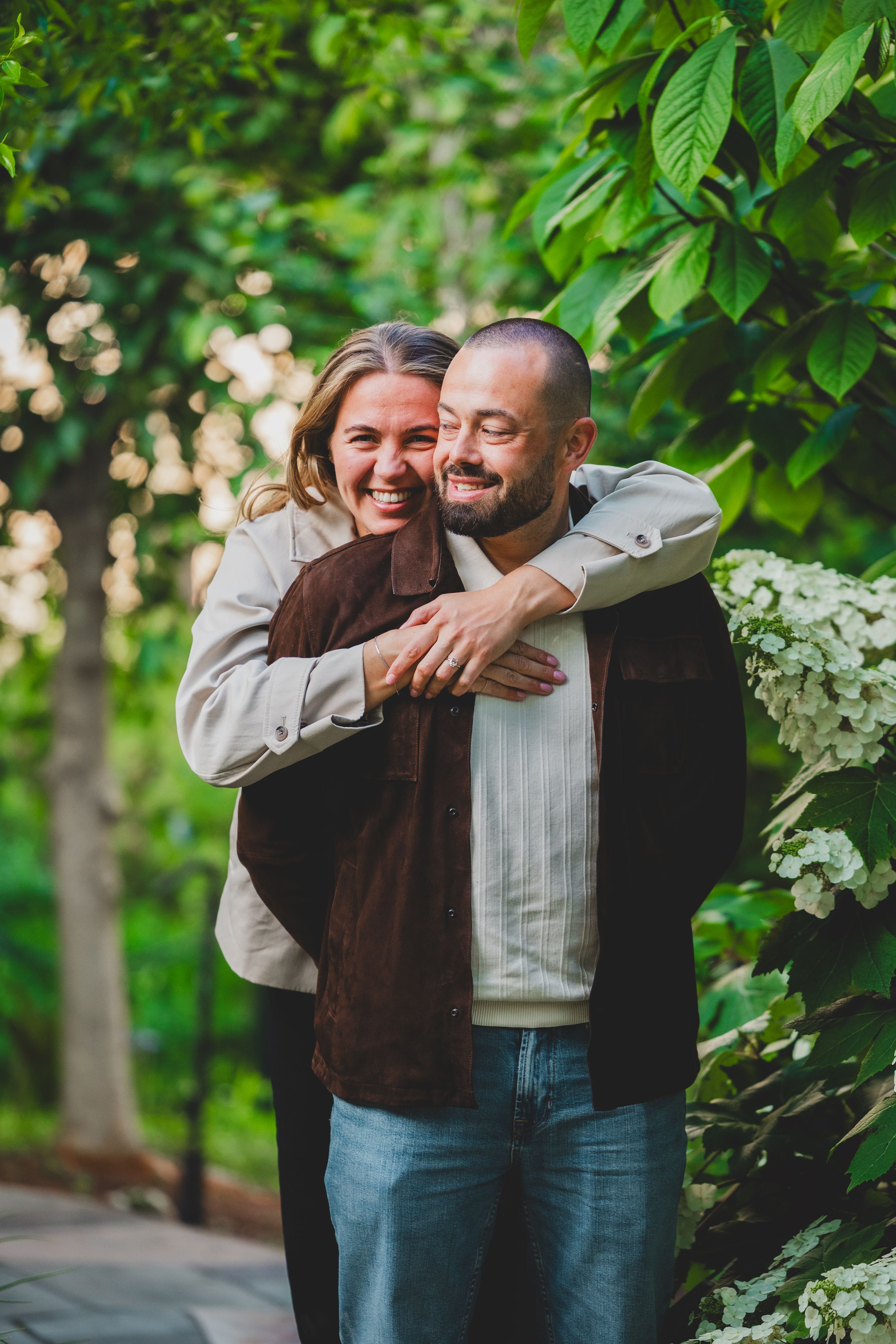A couple embraces and laughs together while standing in a lush green garden setting.