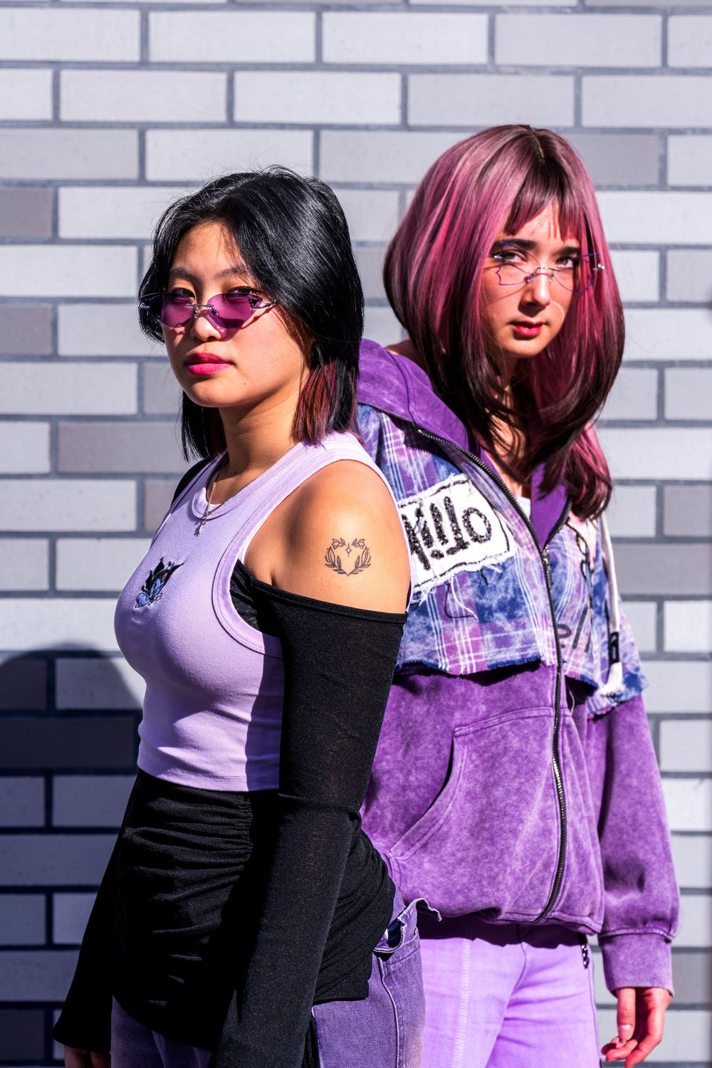 Two people in coordinated purple and black outfits pose against a white brick wall in an urban fashion photoshoot.