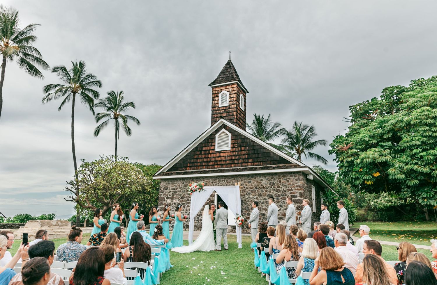 A wedding ceremony takes place at a charming stone chapel in Hawaii with palm trees and guests in turquoise attire.