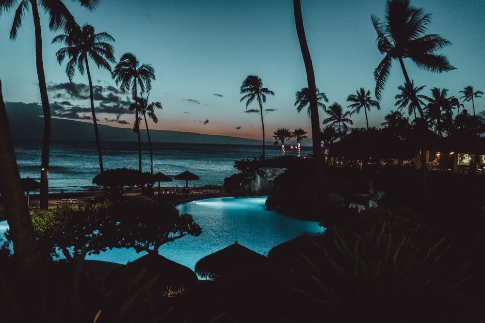 Twilight view of tropical resort with illuminated pool and palm trees silhouetted against dusky sky.