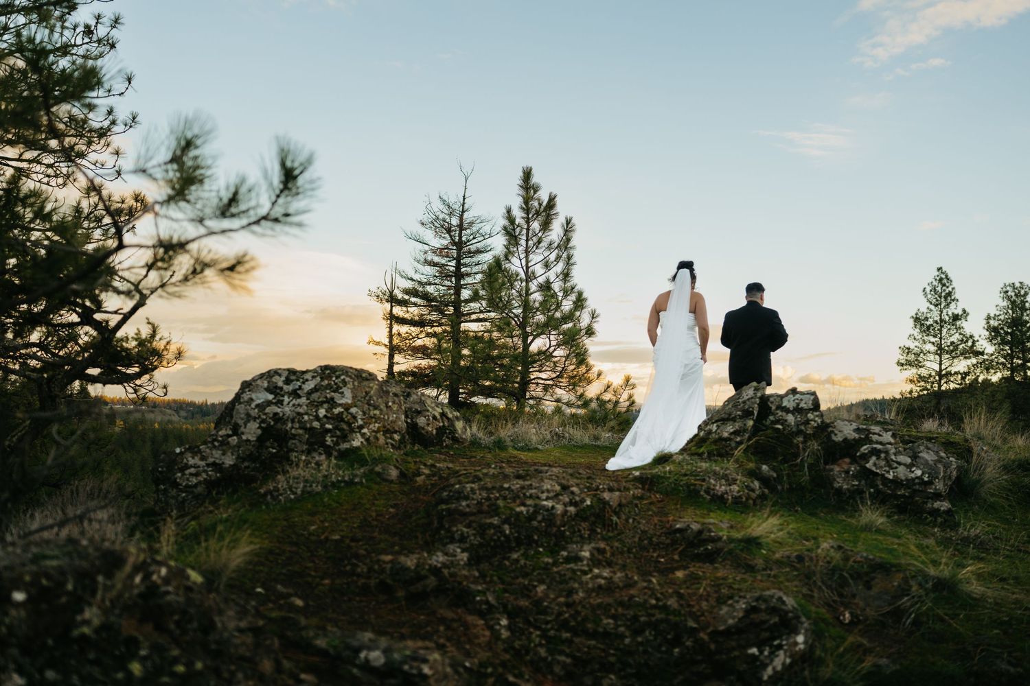 A couple in wedding attire stands silhouetted against a dramatic sunset sky on a rocky mountain ridge.