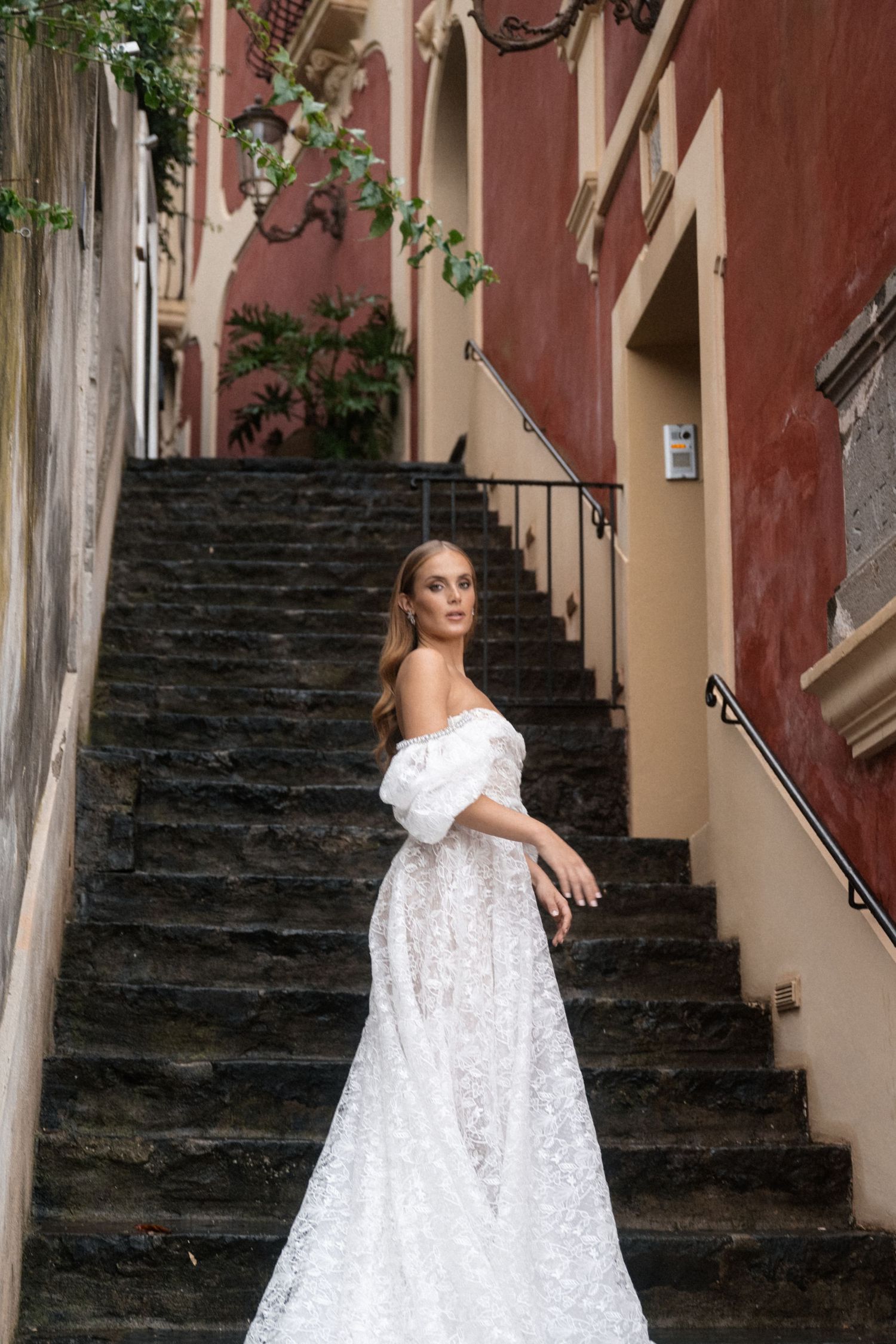 A bride in an elegant lace gown poses on stone steps against a rustic pink wall.