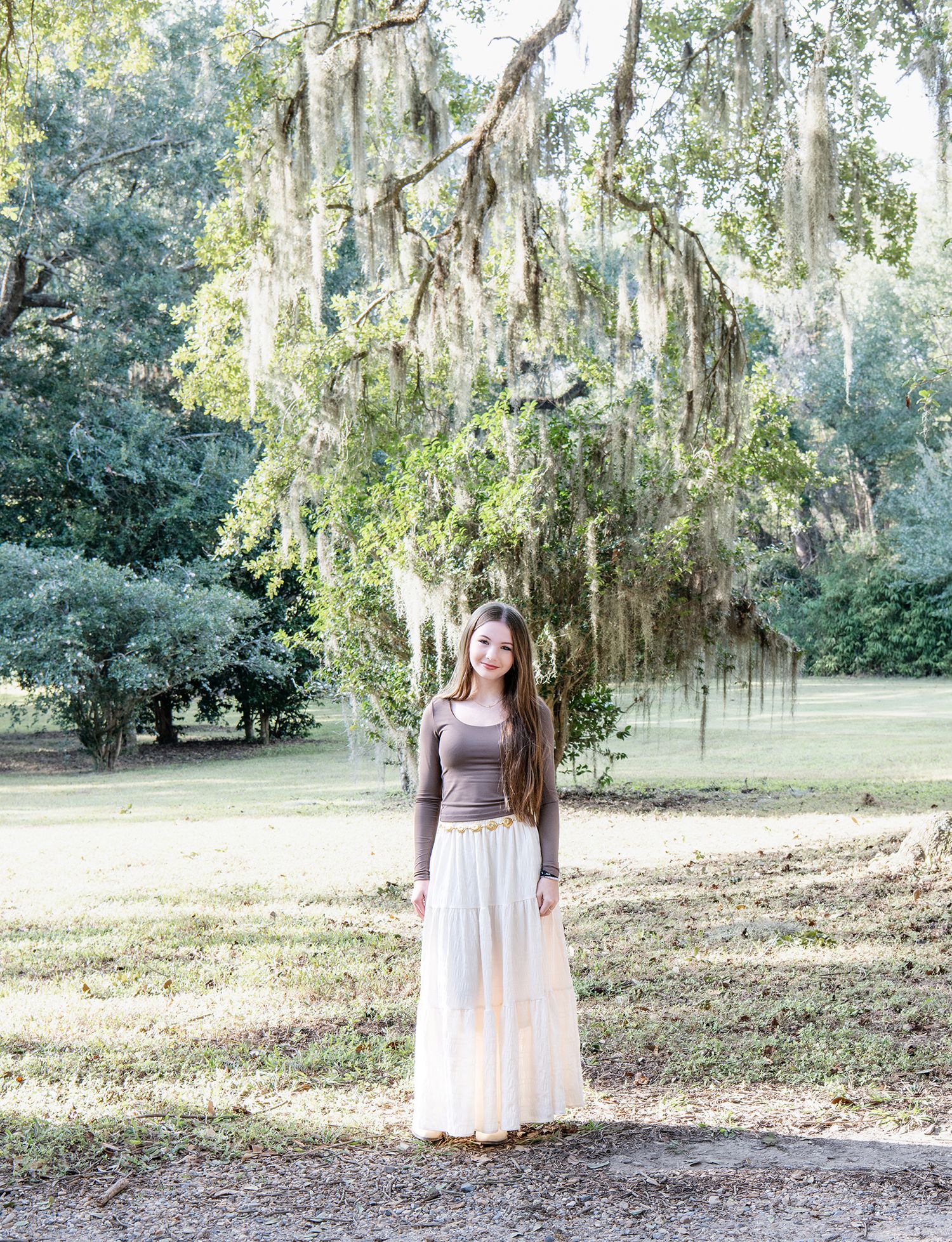 Young woman in white maxi skirt standing under Spanish moss-draped oak tree in a sunlit park.