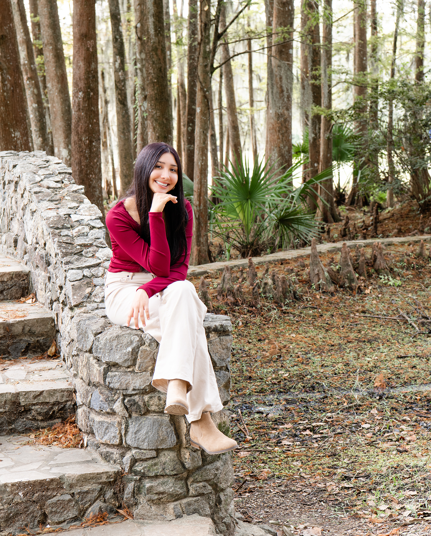 Smiling young woman in red sweater sitting on stone wall steps surrounded by pine forest.