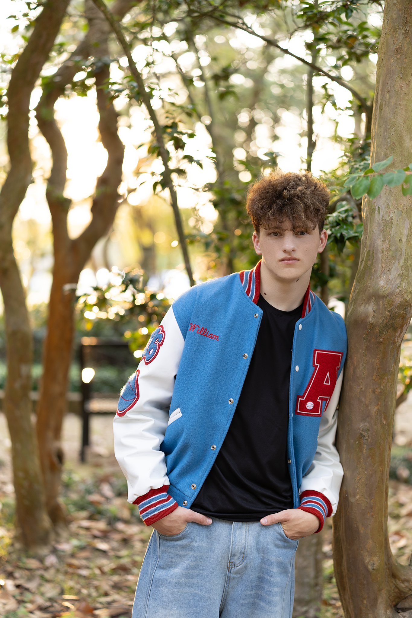 Young man in blue varsity jacket with red accents leans against tree in autumn park setting.