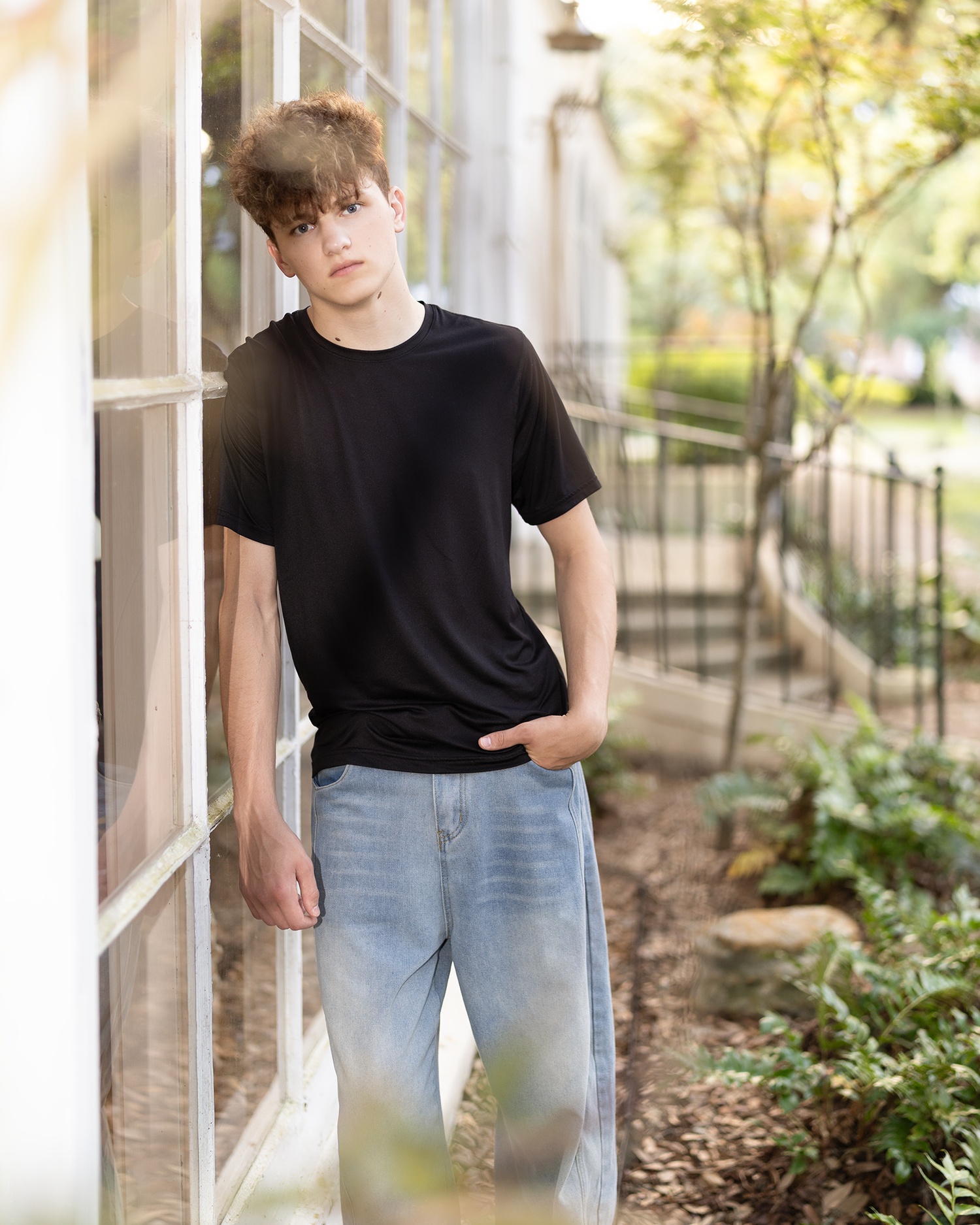 Teen boy in black t-shirt and light jeans leaning against large window in outdoor garden setting.