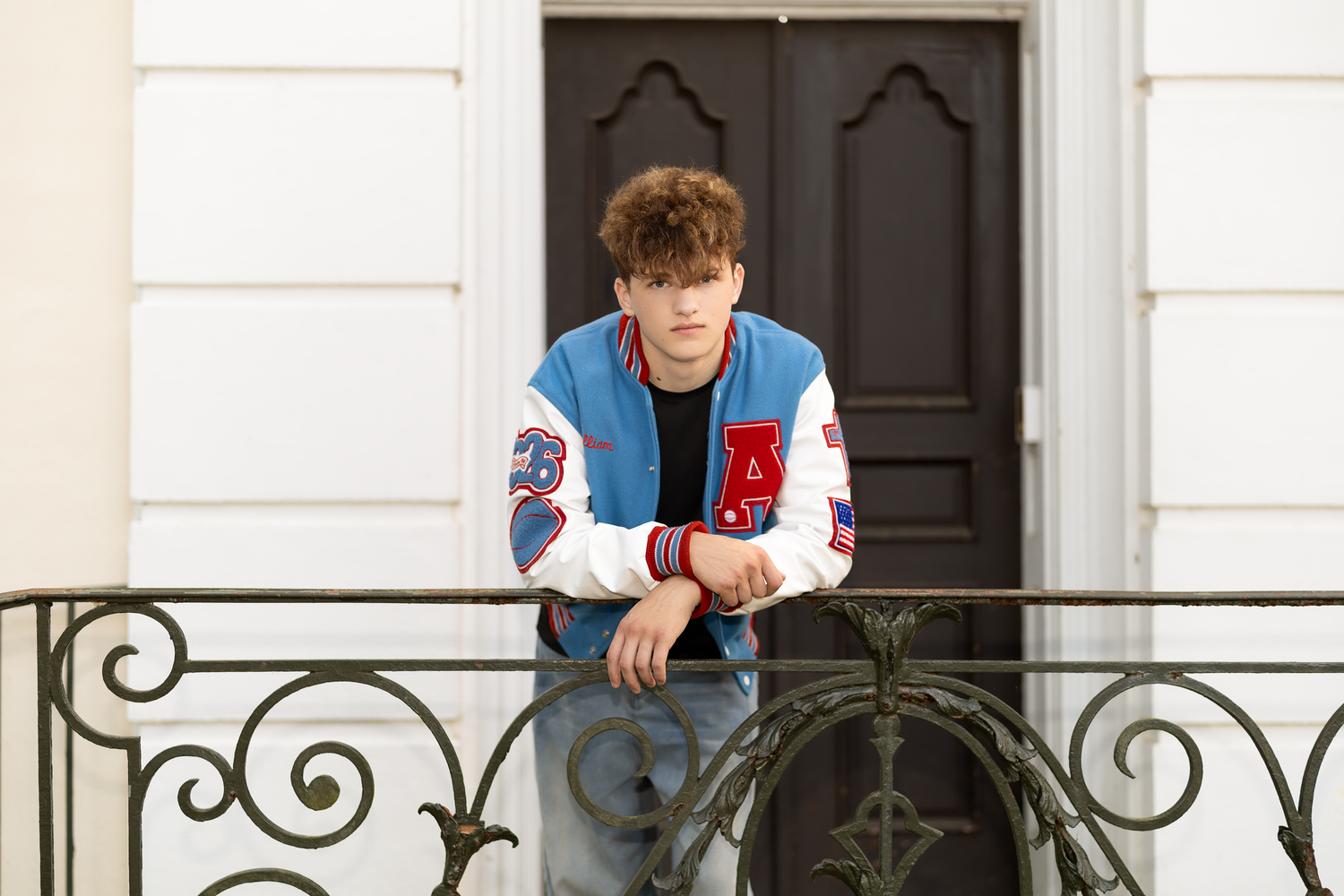 Teen boy in blue and white varsity letterman jacket poses on ornate balcony railing.