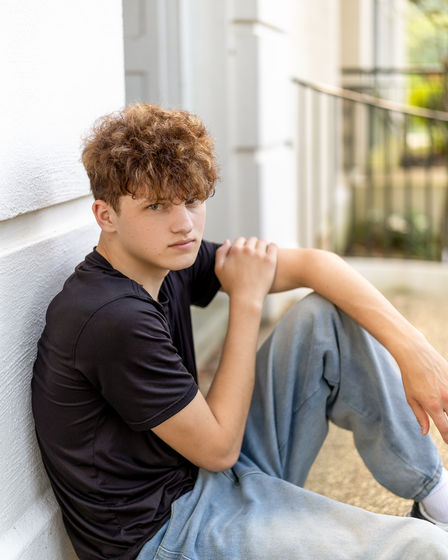 Young man with curly hair in black t-shirt and jeans sitting against white wall in outdoor setting.