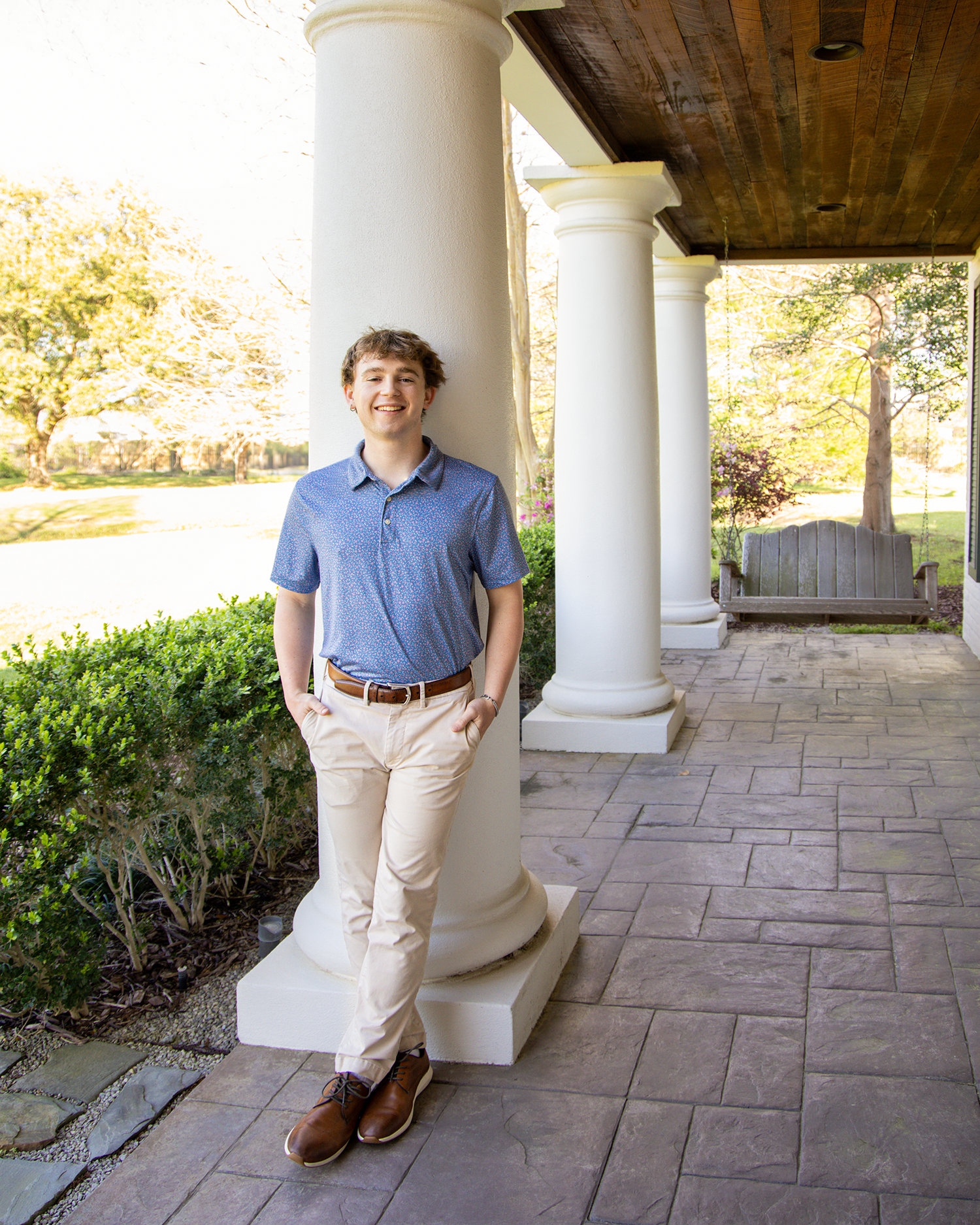 Smiling teen in blue polo and khakis leans against white column on elegant porch.